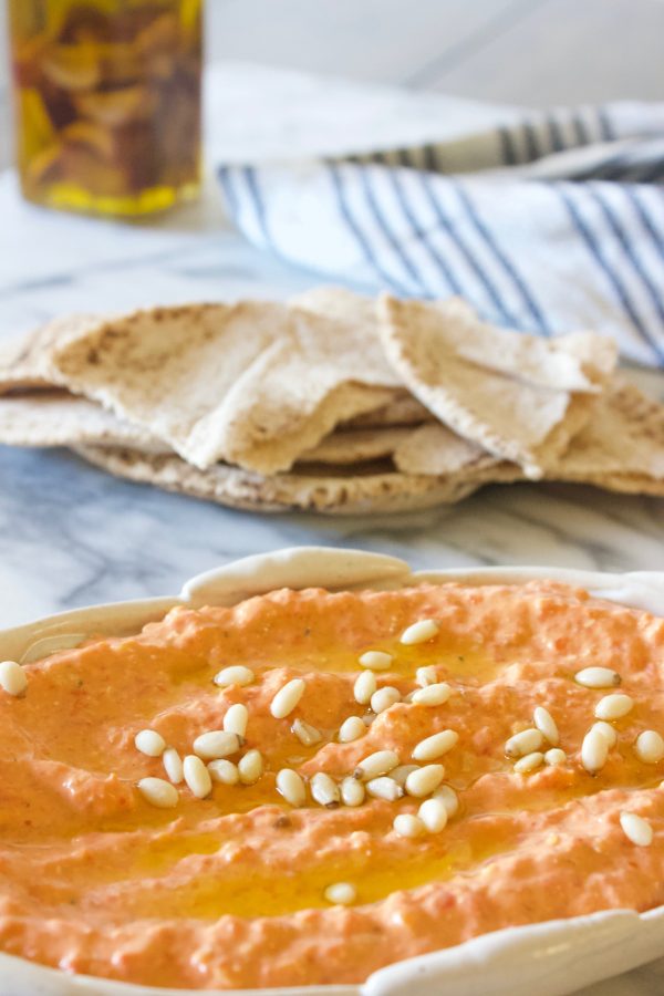Roasted red pepper sauce with pine nuts in a shallow beige dish with on a marble counter with pita and olive oil