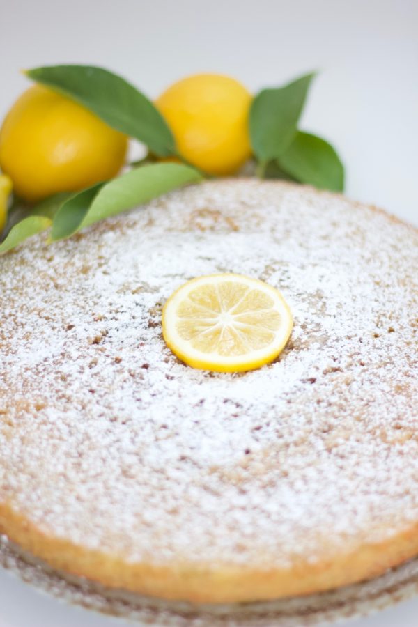 Lemon olive oil cake topped with powdered sugar and a slice of lemon and lemons with leaves in the background