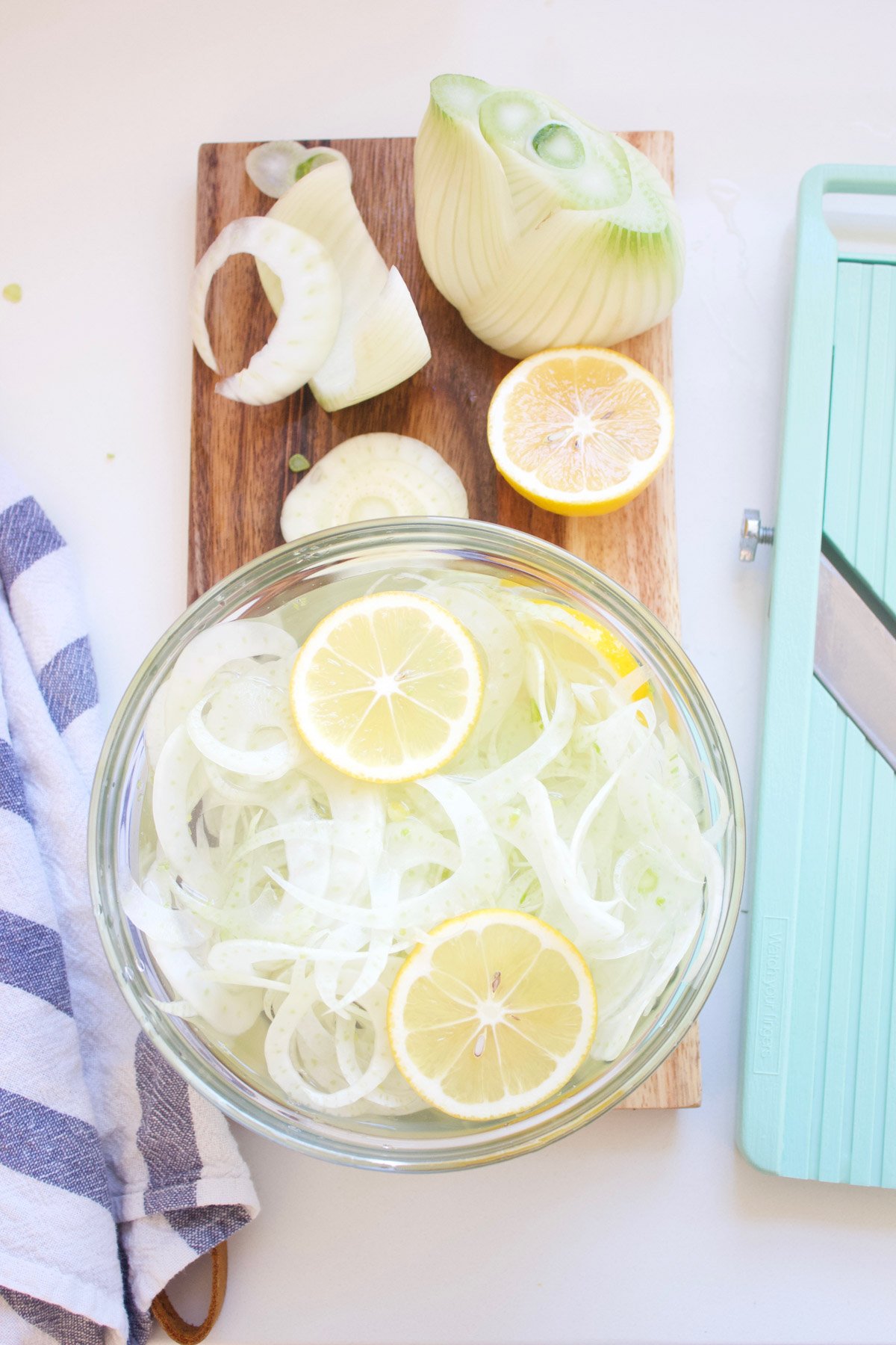 Sliced fennel in a bowl of water with a slice of lemon, sitting on a wooden cutting board.