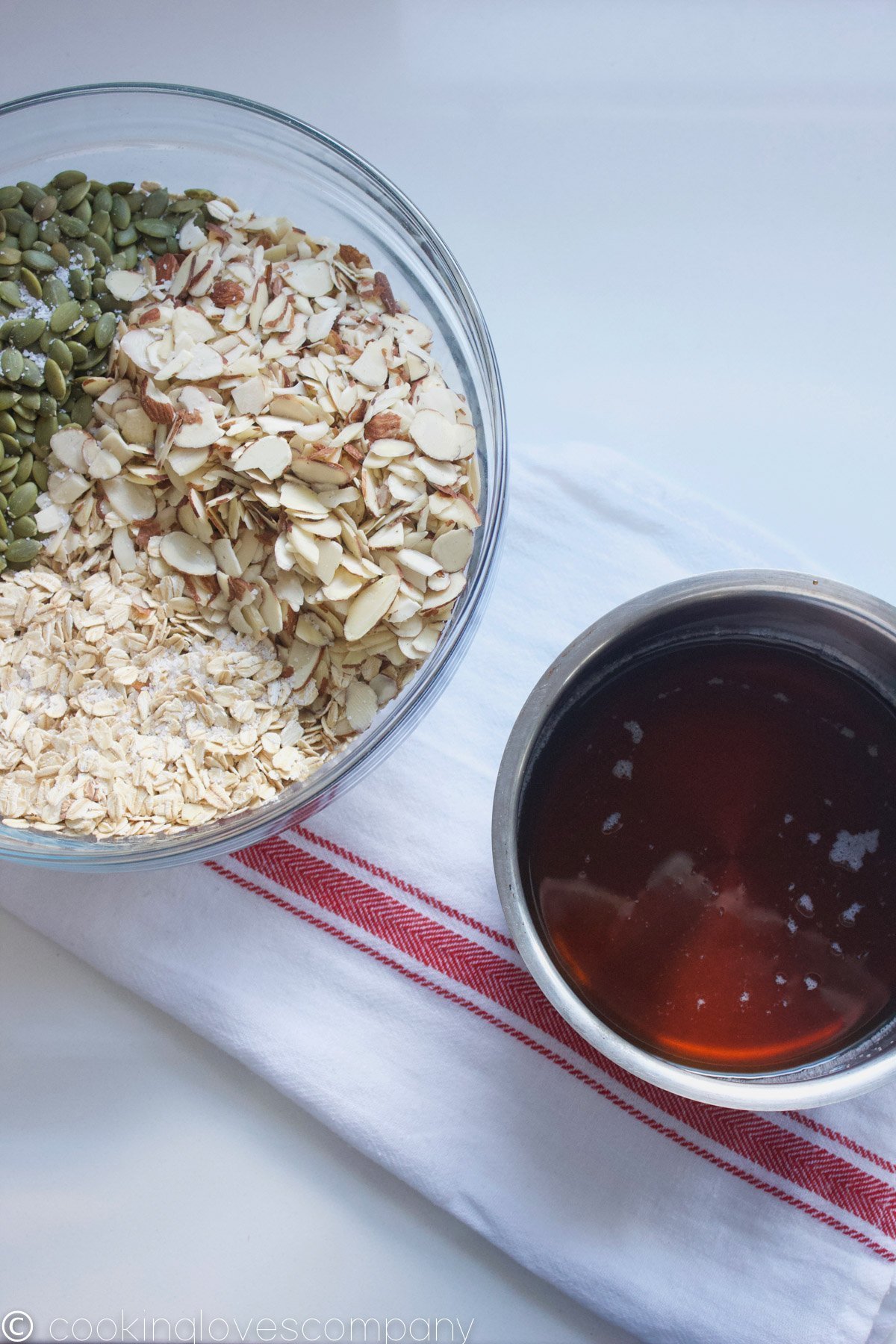 Pumpkin seeds, rolled oats and slivered almonds in a bowl with a small sauce pot with maple syrup and coconut oil on the side