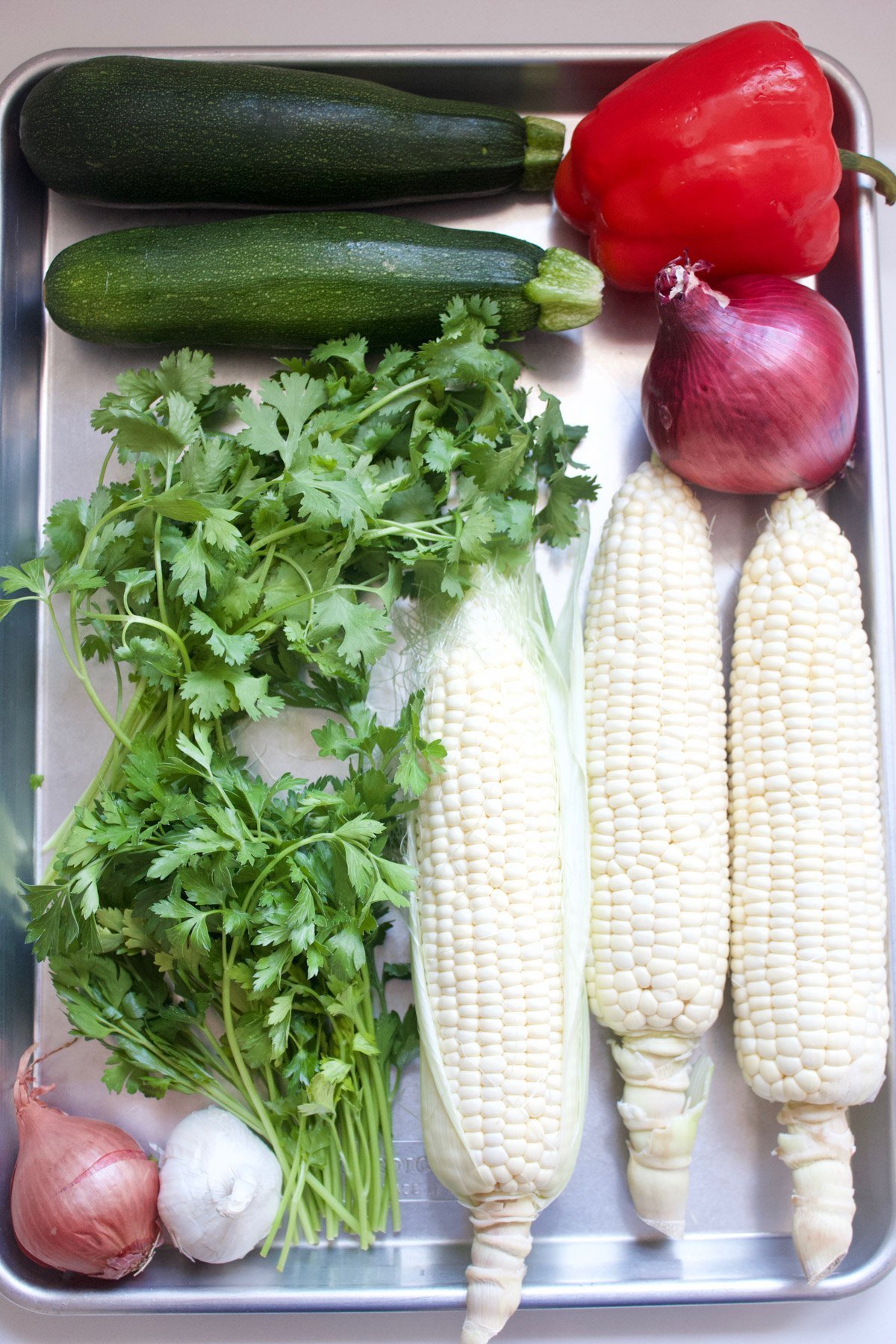 Ears of corn, shallot, garlic, cilantro, red onion, red bell pepper and zucchini on a sheet pan. 