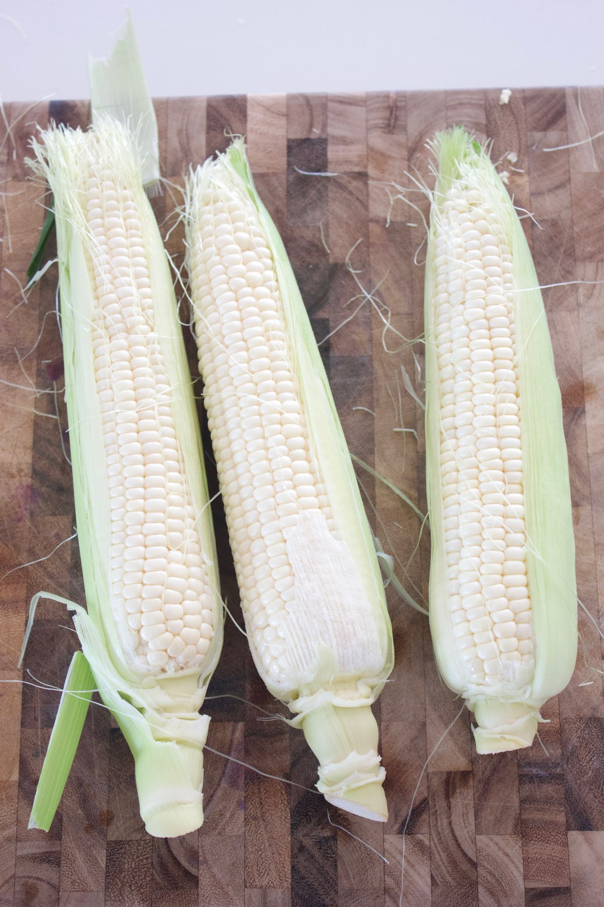 3 ears of partially shucked corn on a cutting board.