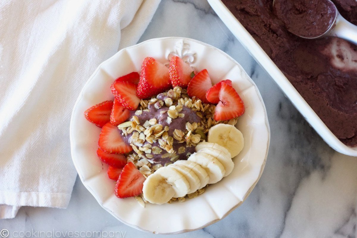 Close up of an assembled acai smoothie bowl in a white bowl on a marble counter with a loaf pan of acai puree on the side and an ice cream scoop in the dish