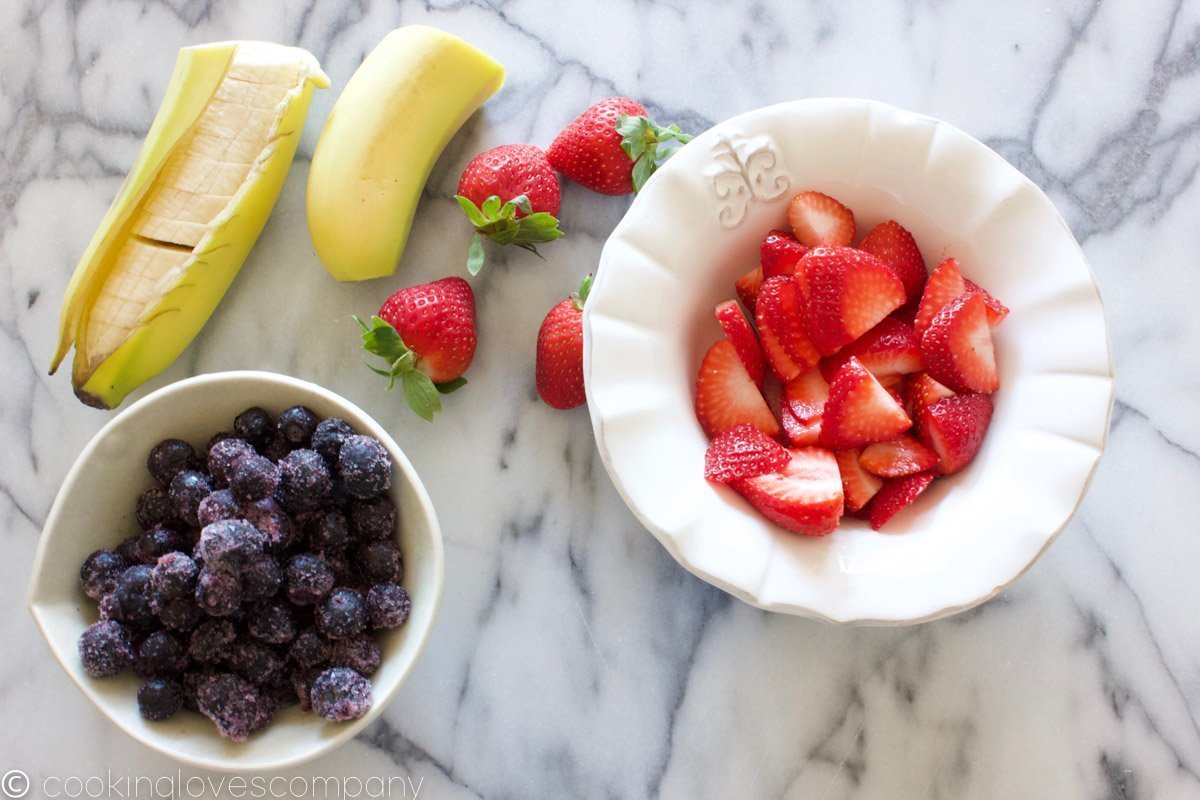 A small bowl of frozen blueberries, a small bowl of strawberries and a banana cut in half on a marble counter
