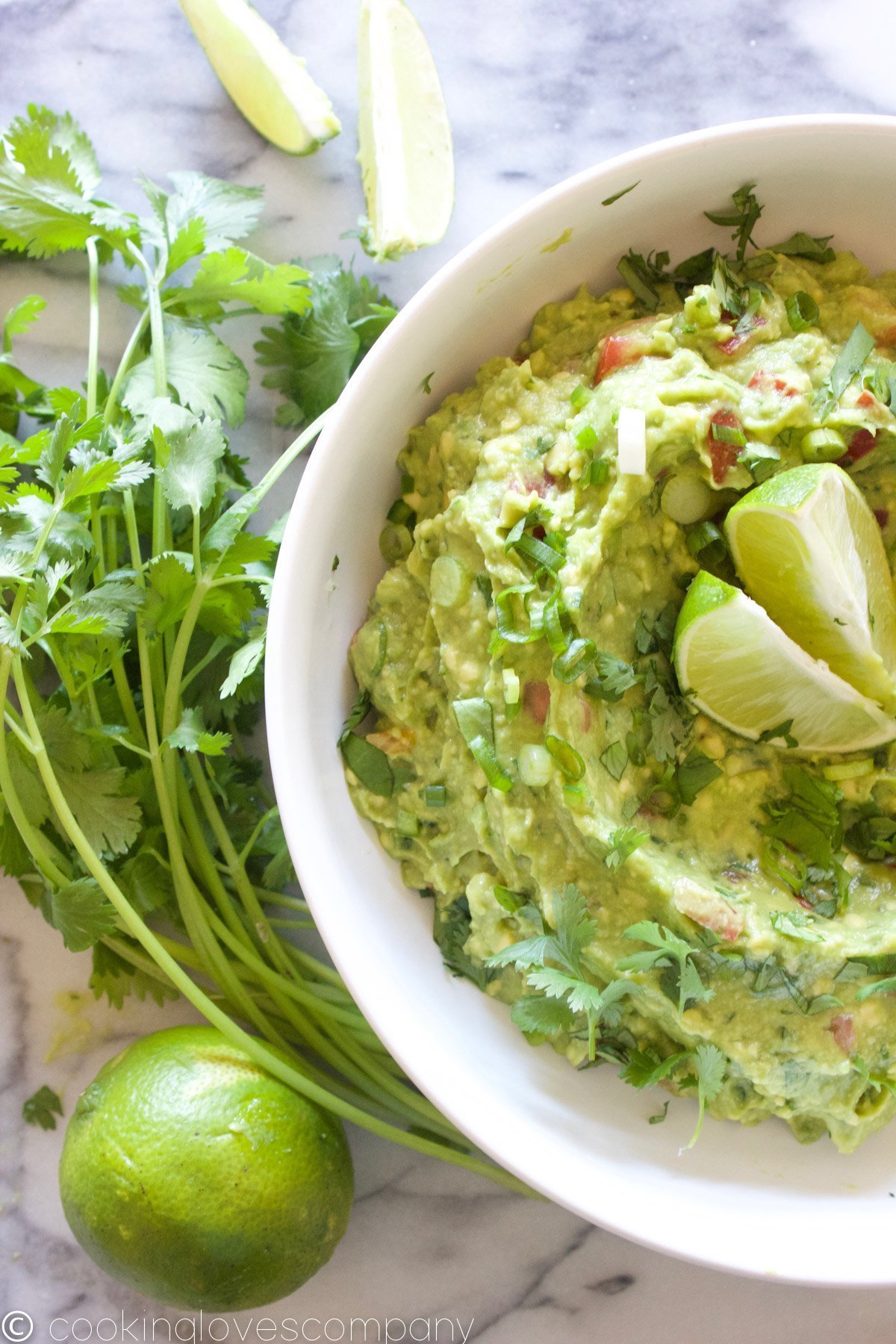 Flatlay of a white bowl filled with guacamole on a marble counter with limes and cilantro on the side 