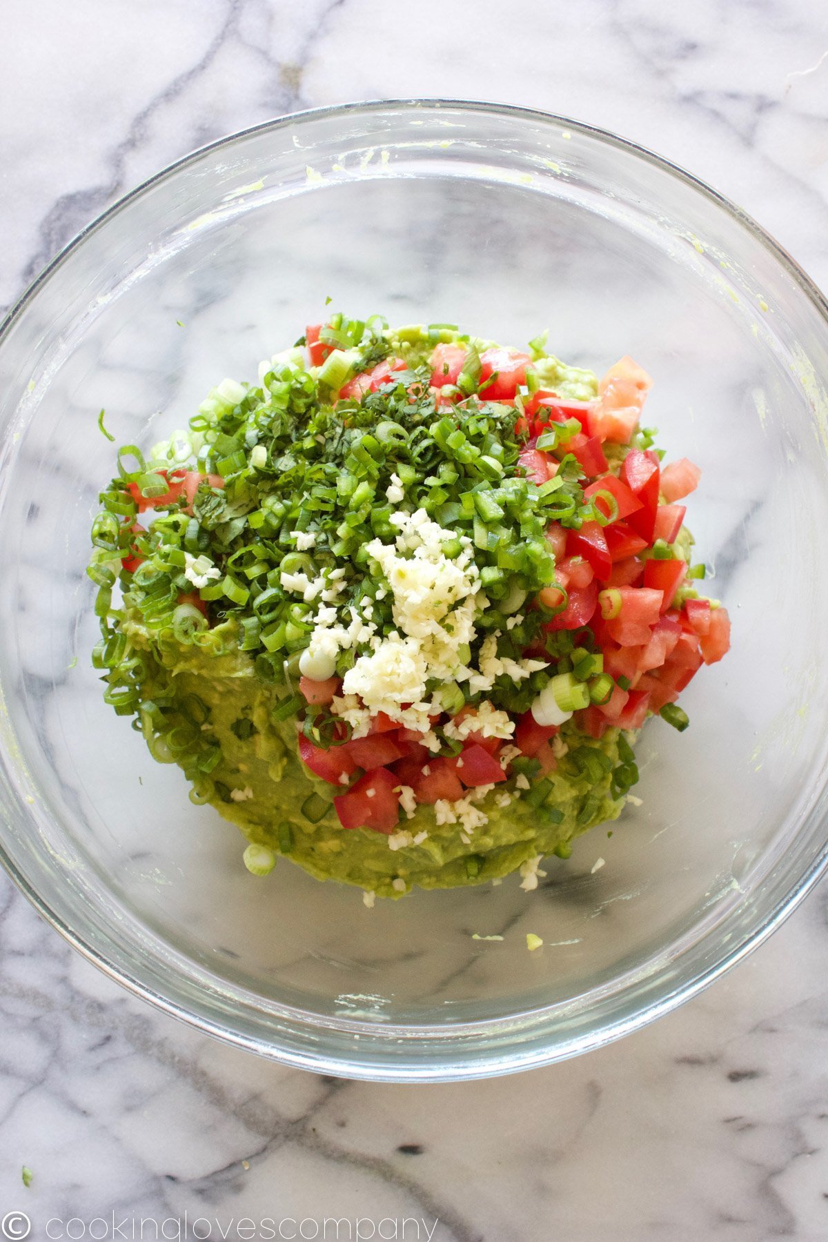 Flatlay of mashed avocado, diced tomato, scallion, garlic, cilantro and jalapeno in a glass bowl on a marble countertop 