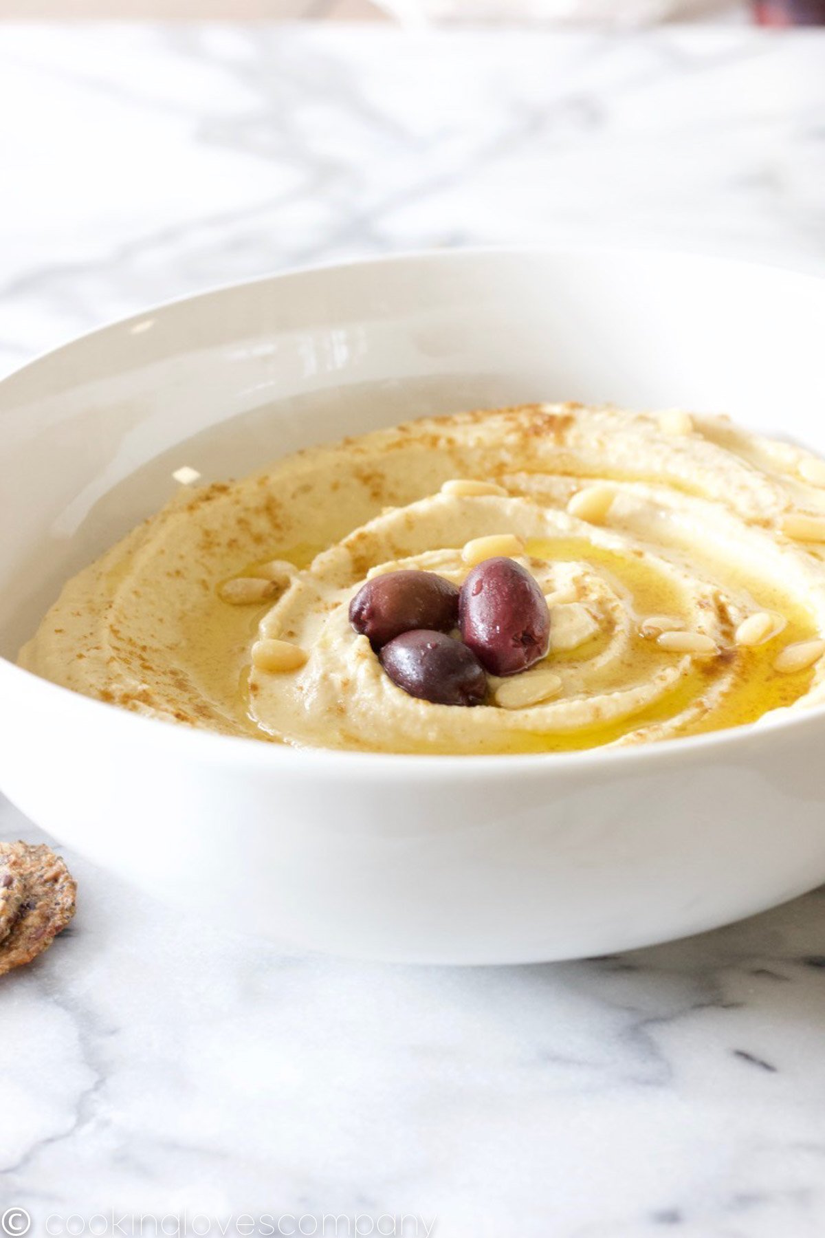 A close up 3/4 angle shot of a white bowl filled with hummus, olive oil, pine nuts and kalamata olives on a marble counter