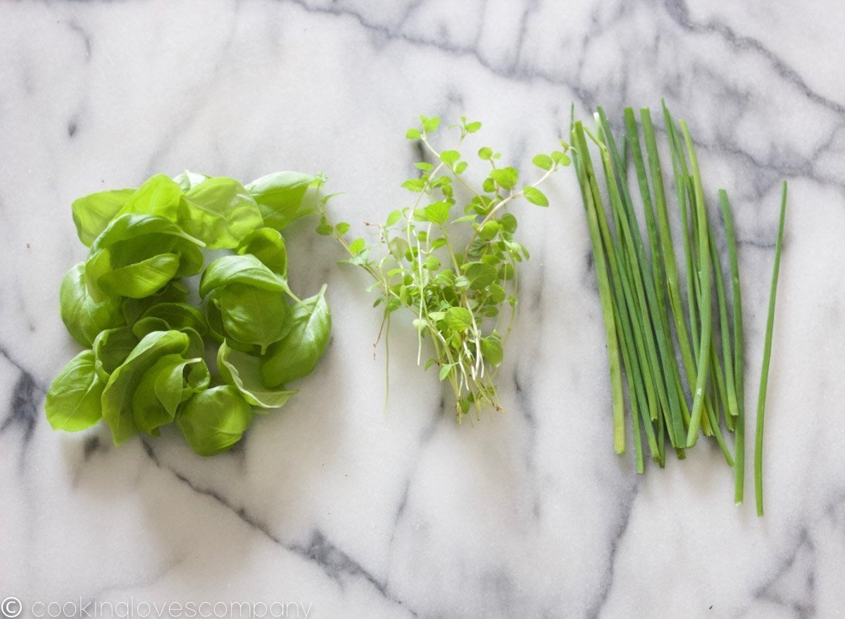 Three separate piles of herbs, basil, mint and chives on a marble counter