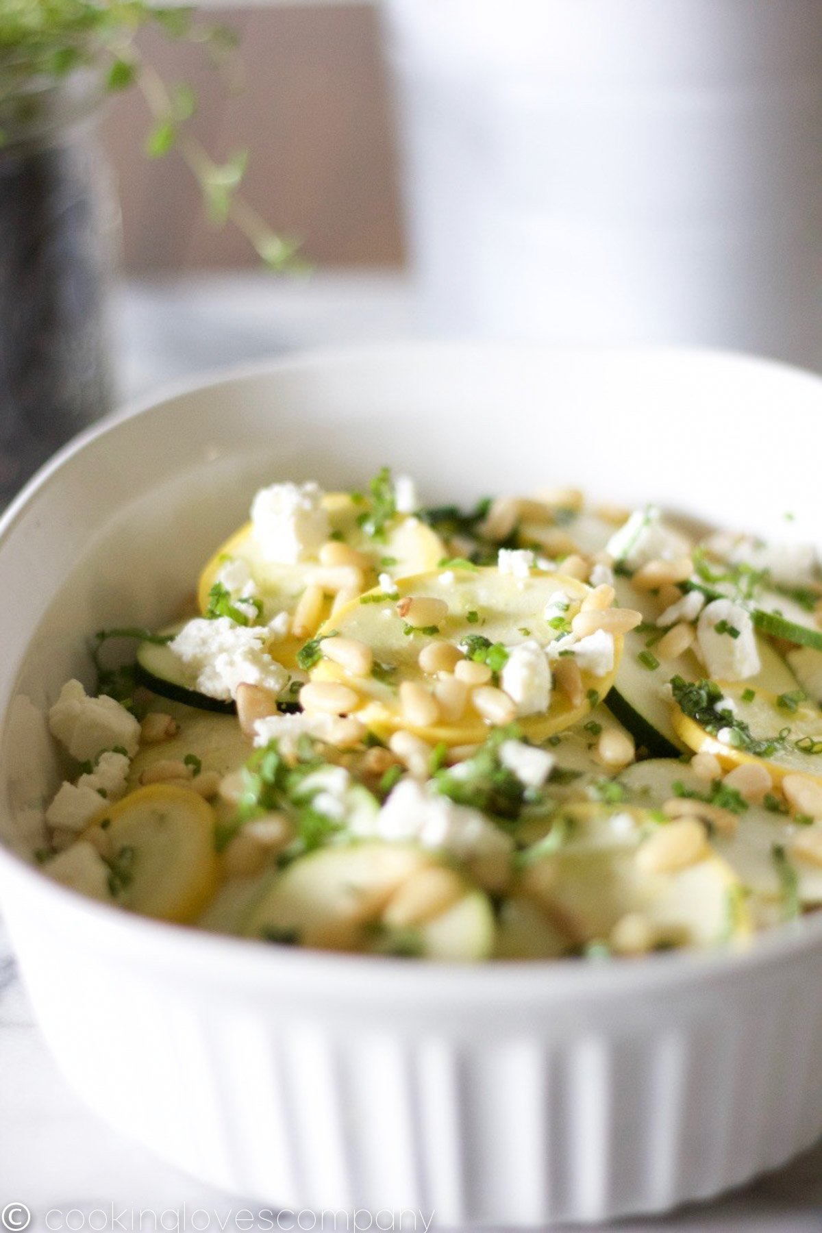Thinly sliced summer squashes with feta cheese, herbs and pine nuts in a white casserole dish