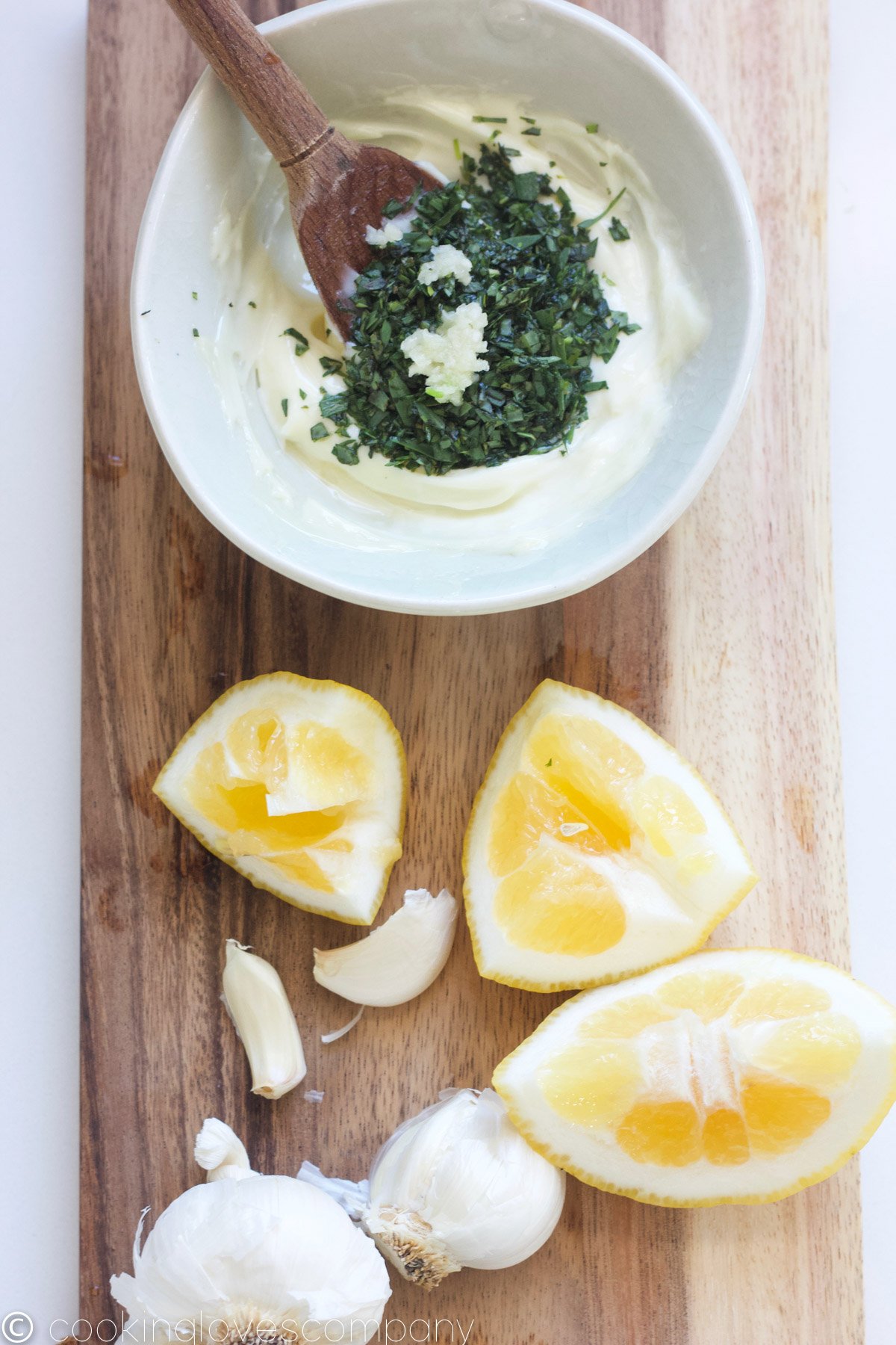 A small bowl of mayonnaise with chopped tarragon and a wooden spoon on a wood cutting board with lemon slices and garlic on the side.