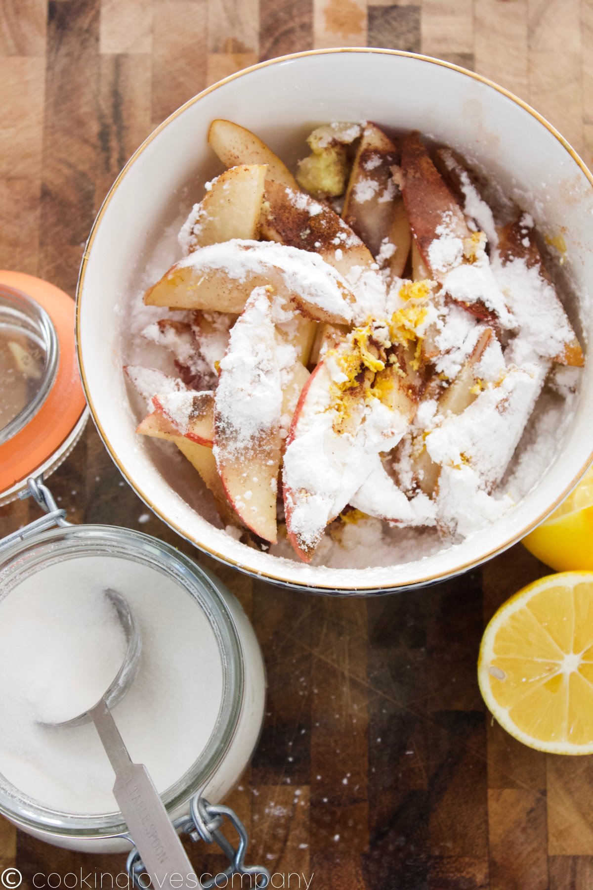 Flat lay of a white bowl with a gold rim filled with apples, pears, flour, ginger and sugar with a halved lemon and bowl of sugar on a cutting board