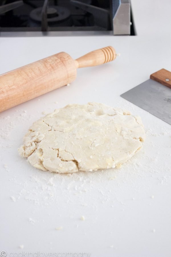 Flattened wheel of pastry dough on a white counter with a pastry scraper and rolling pin in the background