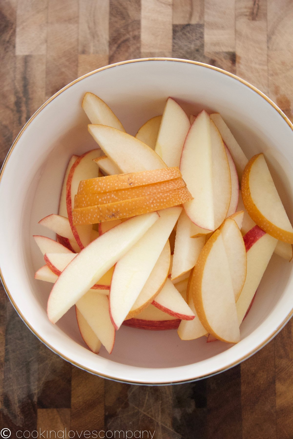 A white bowl with a gold rim on a wooden cutting board filled with sliced apples and pears